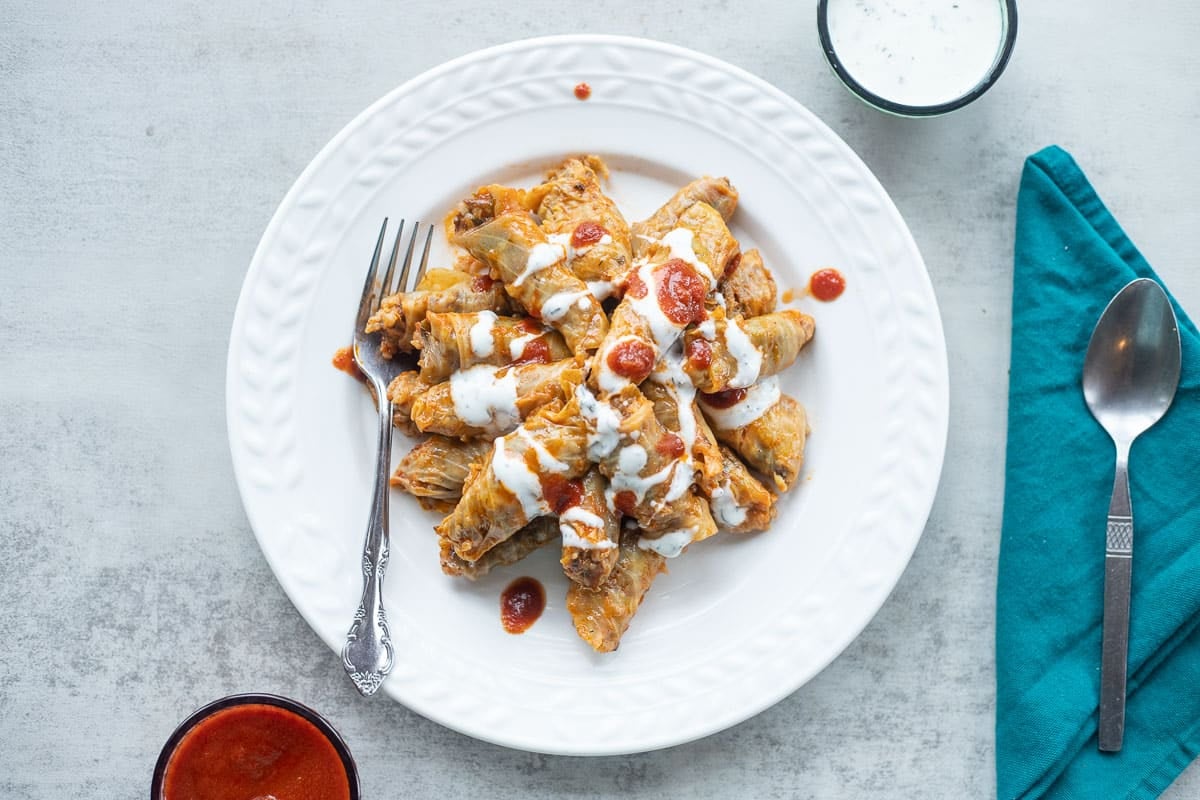 Assyrian cabbage dolma on a white plate with  a folded blue napkin and red and white sauce on the side.