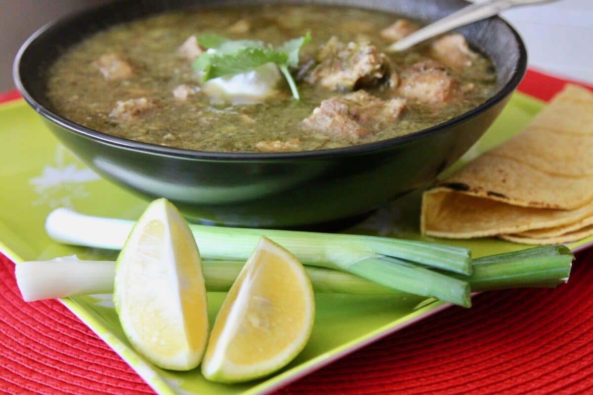 chili verde in a dark bowl with green onions, lime wedges and tortillas on the side