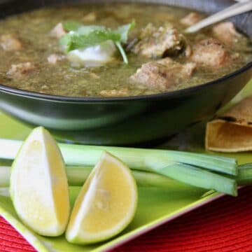 chili verde in a dark bowl with green onions, lime wedges and tortillas on the side