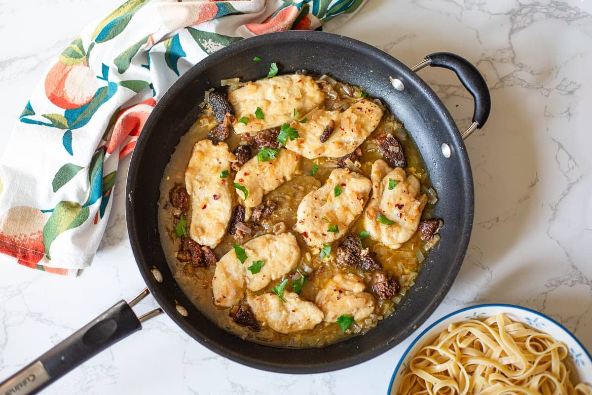 chicken scallopini in a skillet with pasta on one side and a colorful dish towel on the other side