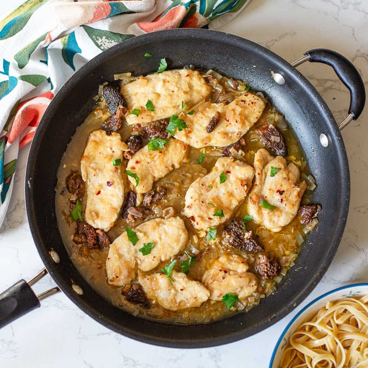 chicken scallopini in a pan with partially visible bowl of past and a colorful dish towel