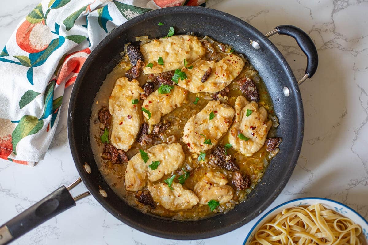 chicken scallopini with morel mushrooms in a large skillet with pasta on the side and a colorful dishtowel next to it