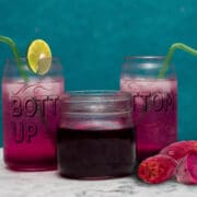 prickly Pear Juice in two glasses with prickly pear syrup in a jar and a sliced prickly pear in front