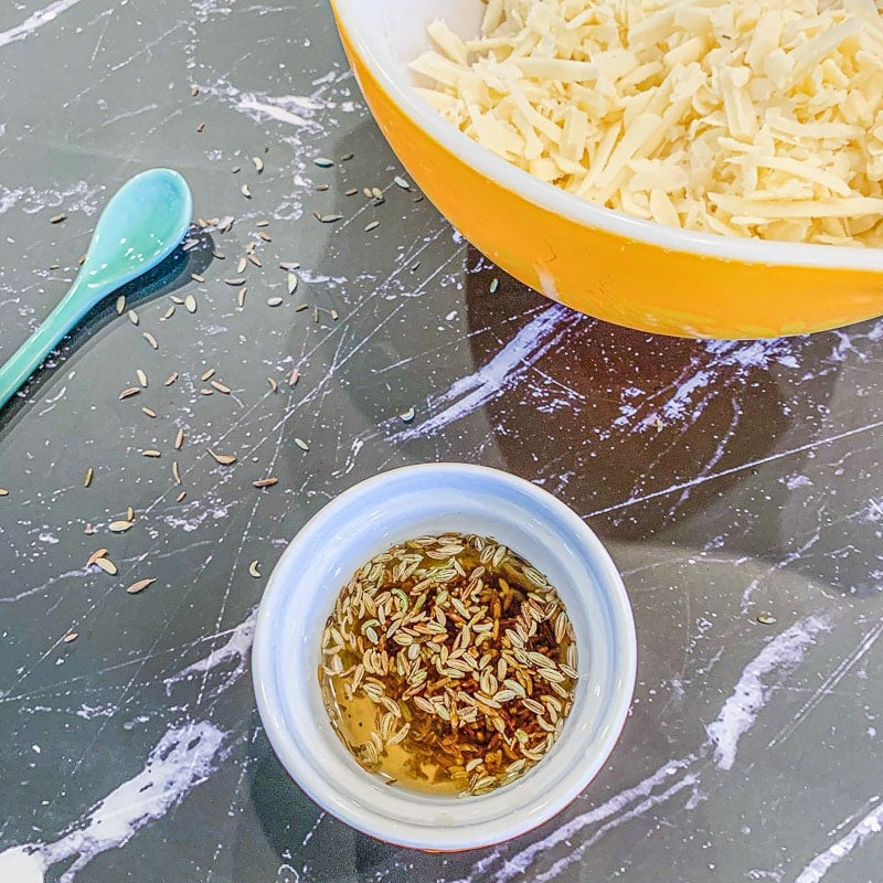 caraway and cumin seeds in a small bow covered with water, with a bowl of grated white cheese in the background