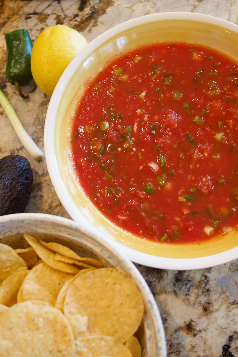 mexican salsa in a yellow bowl with tortilla chips in a bowl next to it and lemon, pepper, and scallion on the counter