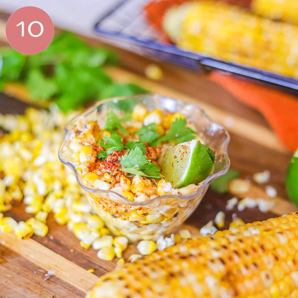 corn on the cob in a plastic cup with corn on the cob, cilantro, and cheese scattered around it