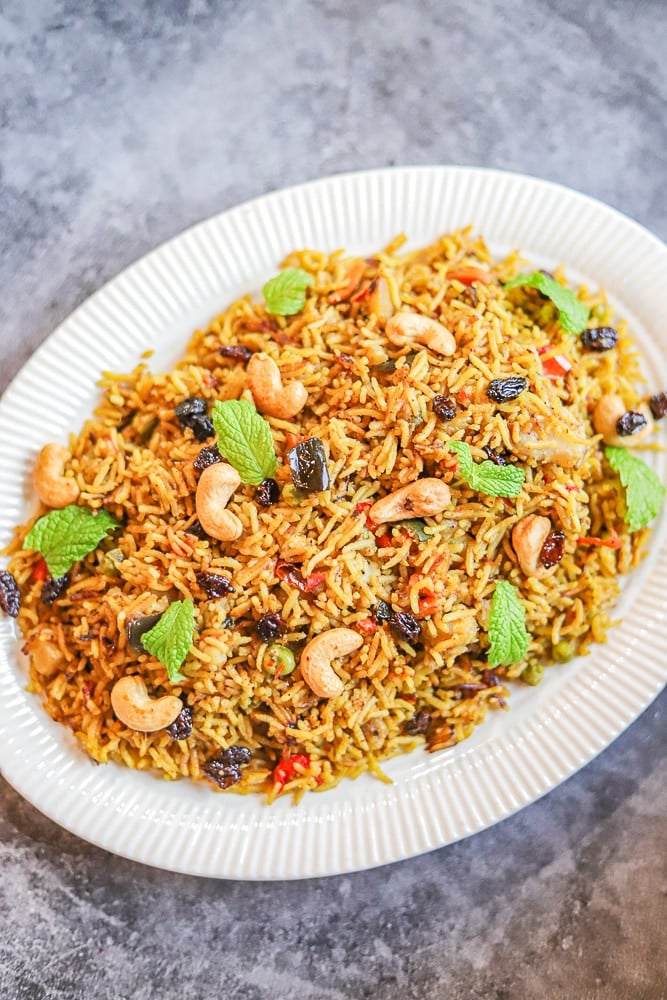 a white plate with vegetable biryani, topped with cashews, raisins, and mint leaves