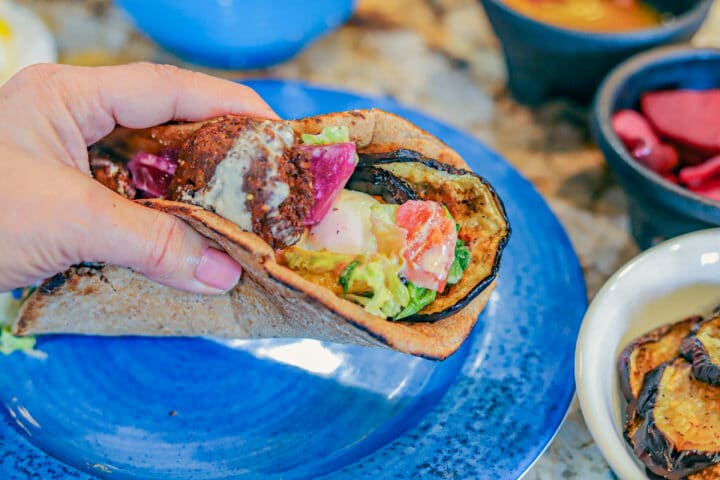 Sabich Sandwich being held over a blue plate with fixings around it