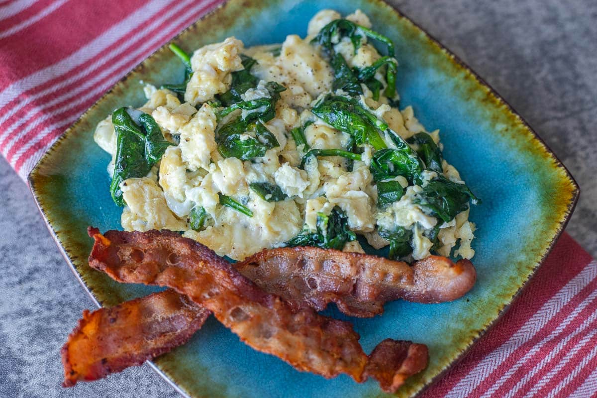 spinach and eggs on a blue plate with 2 strips of bacon