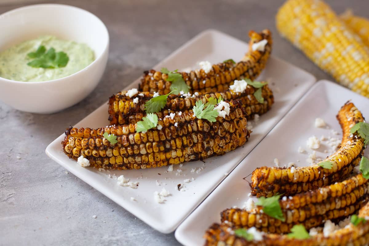 corn ribs topped with cotija cheese and chopped cilantro with cilantro lime sauce and fresh corn in background