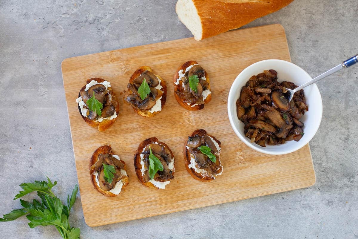 mushroom bruschetta with goat cheese and parsley to garnish, all on cutting board beside baguette