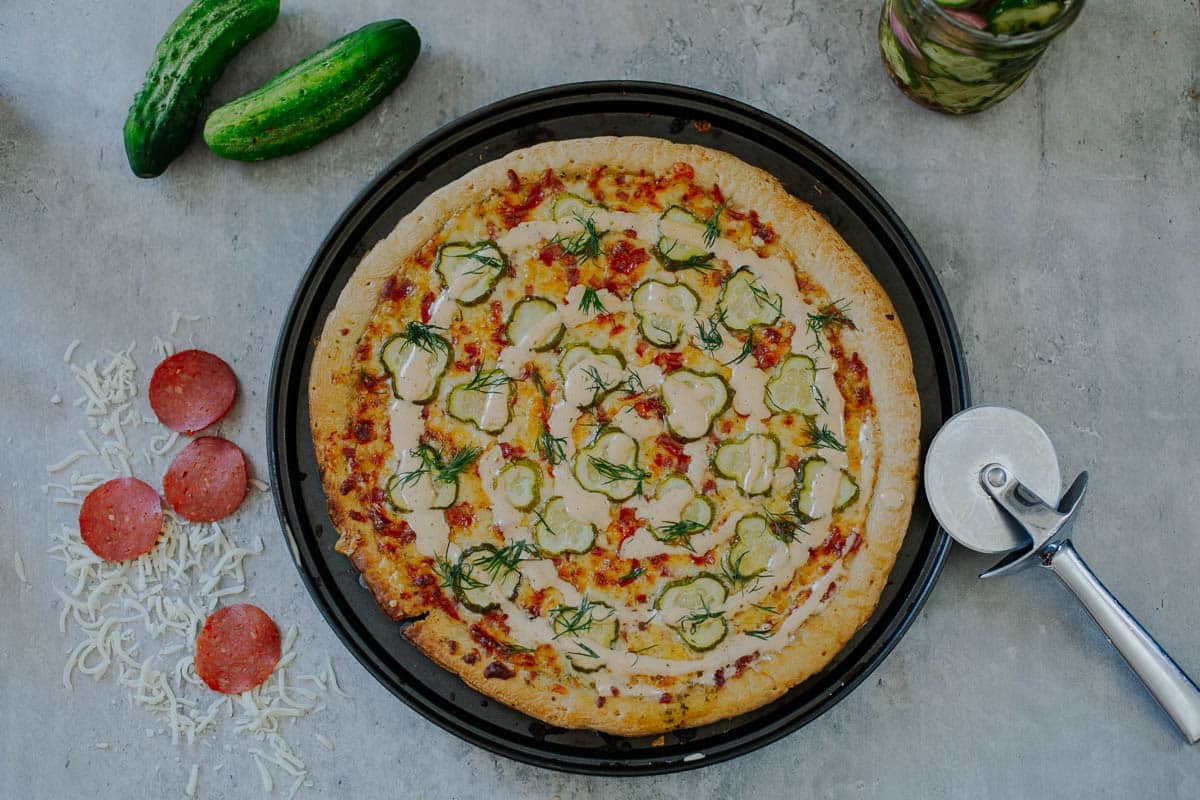 dill pickle pizza surrounded by cheese, pepperoni, cucumbers, pizza cutter