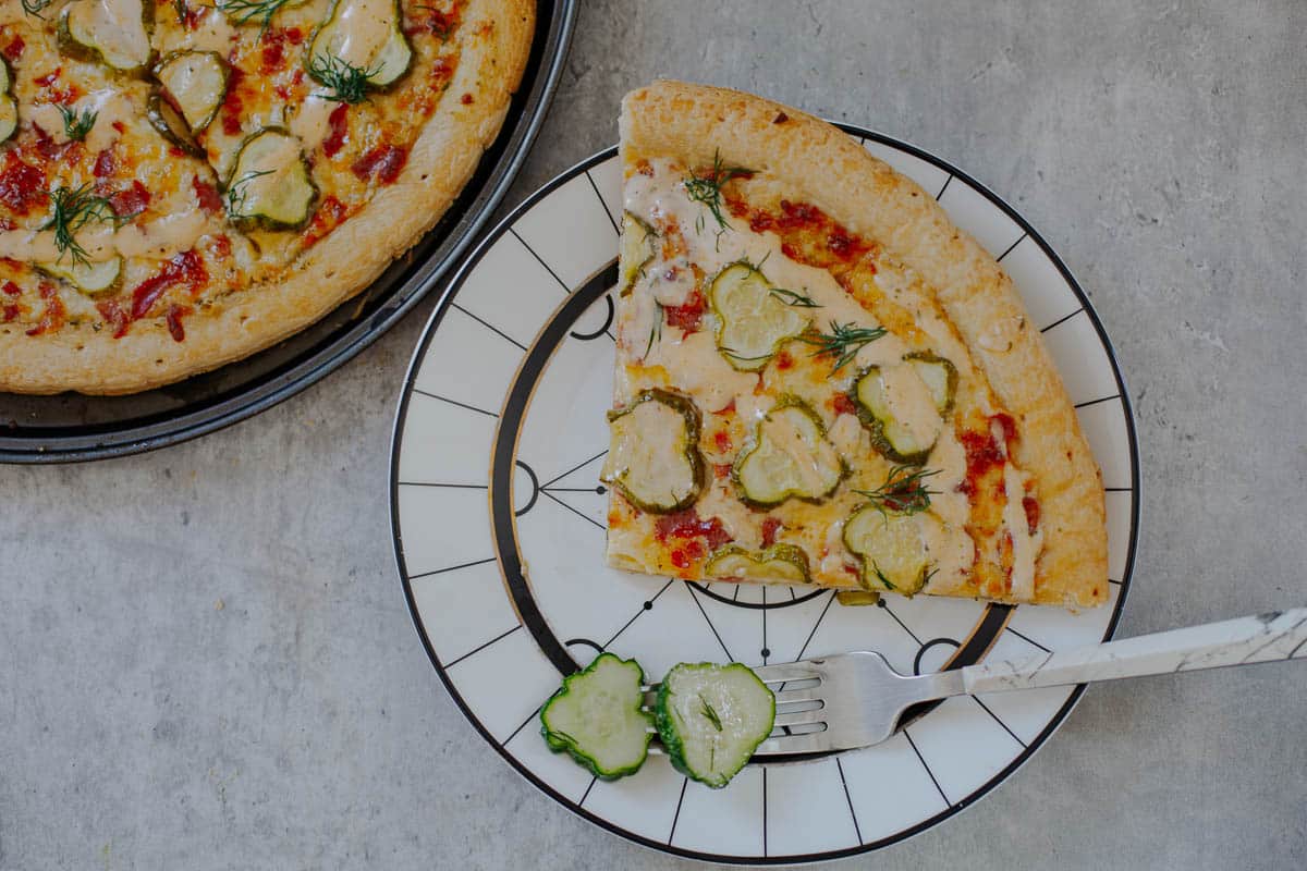 slice of dill pickle pizza on plate with fork and pickles slices beside remaining pizza