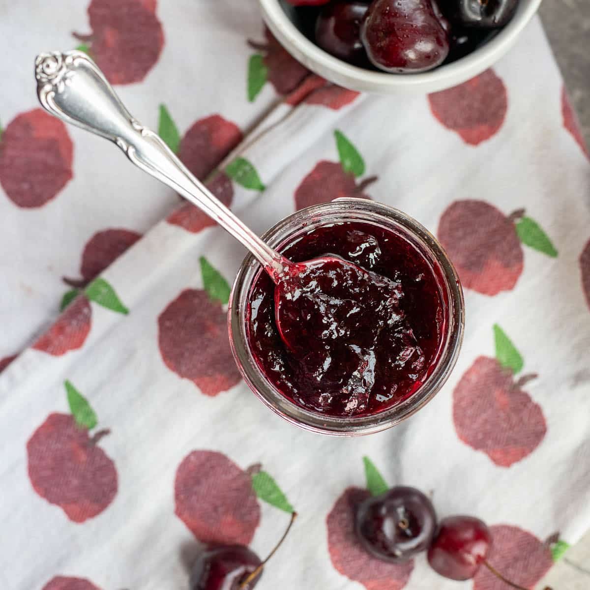 cherry jam in a jar with a spoon in it on a dishcloth with cherries around it