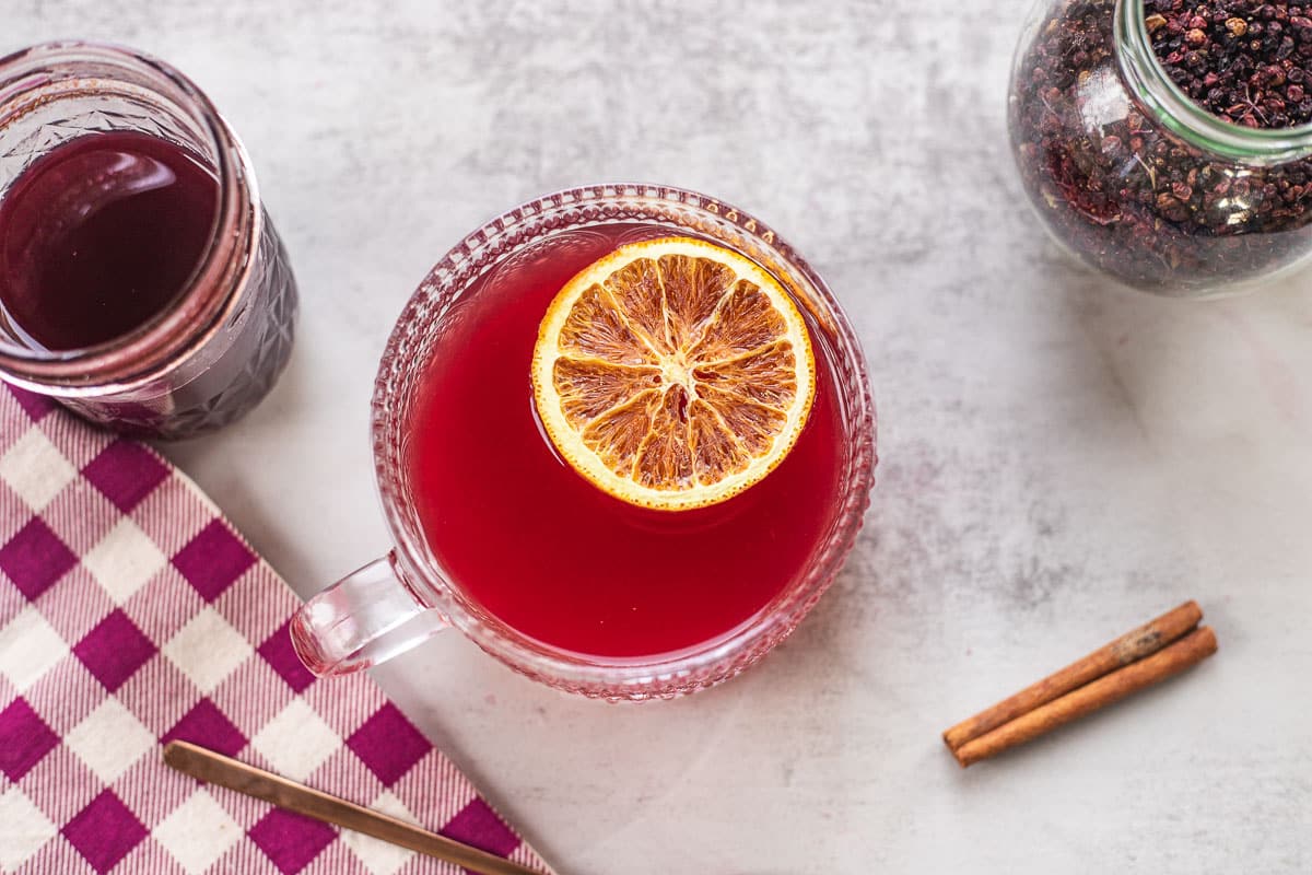 elderberry tea in glass teacup with cinnamon stick, flannel, elderberry syrup, and jar of elderberries
