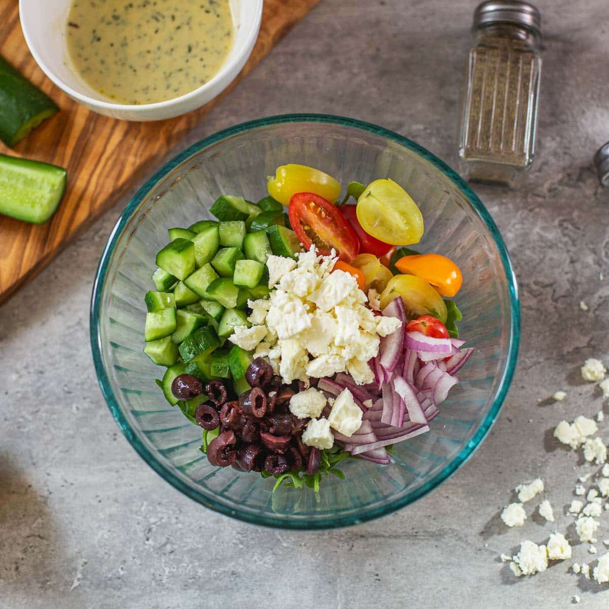 greek cucumber salad in glass bowl before mixing, dressing in bowl on the side along with cucumbers, pepper shaker, feta cheese