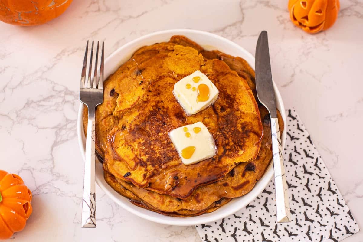 pumpkin spiced pancakes on plate with butter, fork, knife, and napkin with bats on it beside small pumpkins