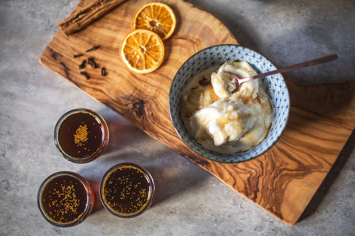 fig syrup in 3 mason jars beside cutting board with cloves, cinnamon stick, orange slices, and bowl of ice cream with syrup on top