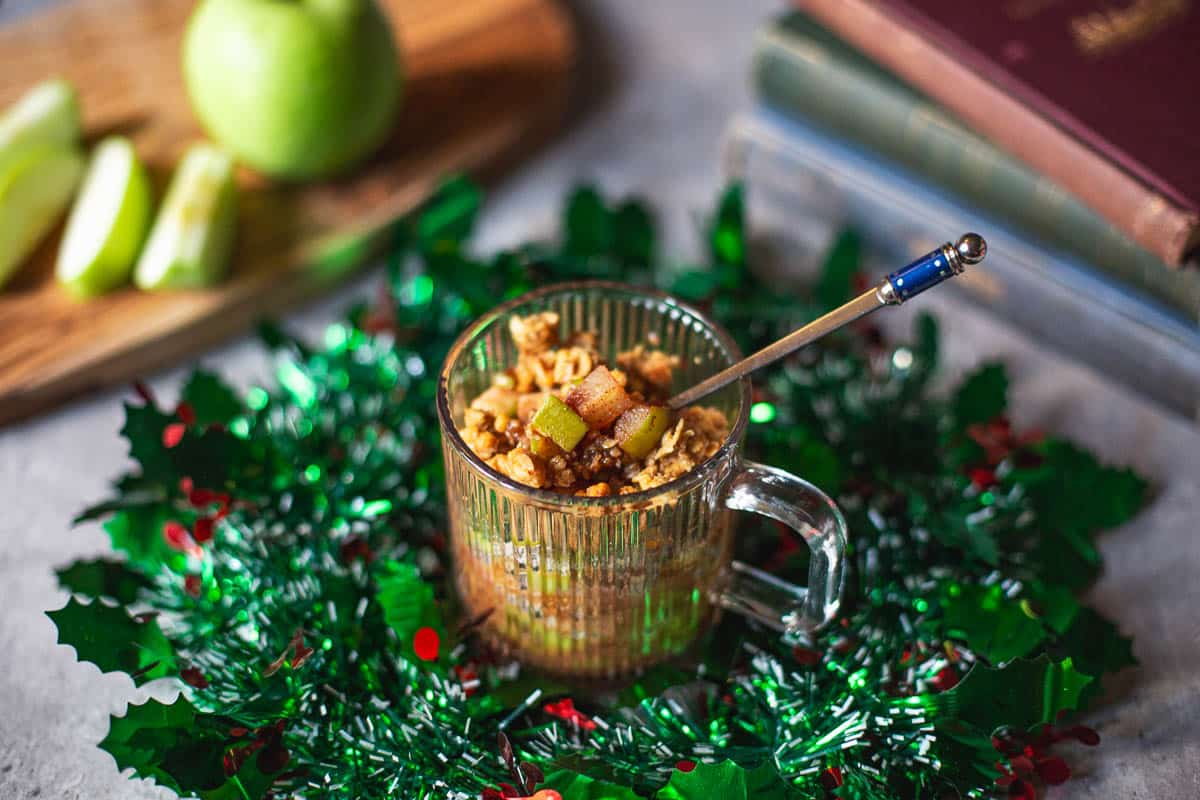 microwave apple crisp in a mug surrounded by wreath with sliced apples and stacked books in background