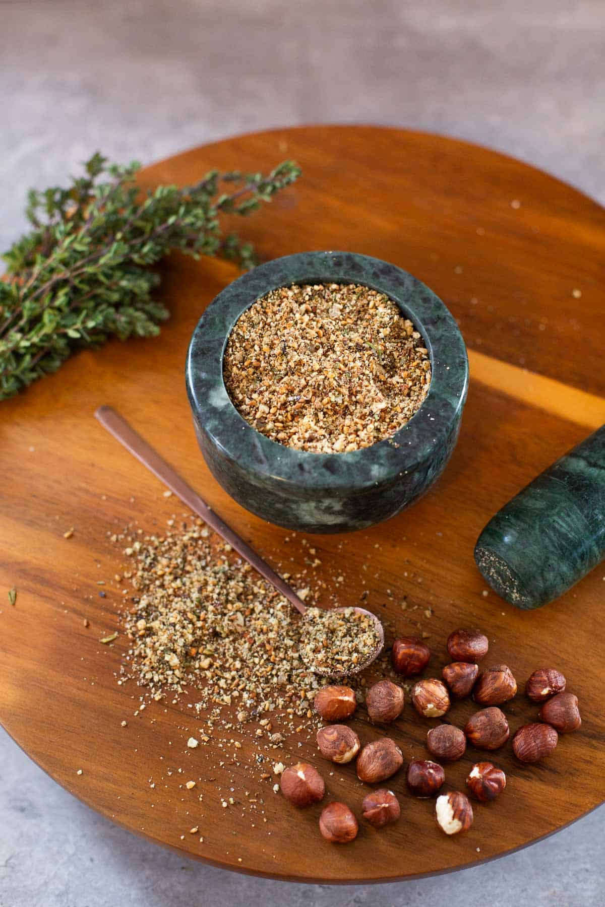 dukka spice in mortar with some spilled on cutting board beside thyme, hazelnuts, and pestle