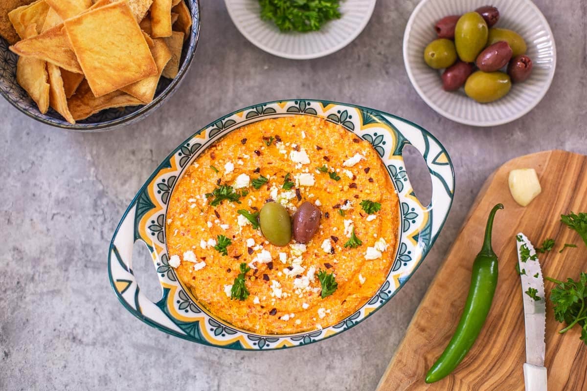 a bowl of tirokafteri (htipiti) beside bowsl with pita chips, olives, chopped parsley, and cutting board