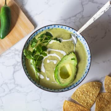 cream of jalapeno soup with avocado half and cilantro bunch on top beside tortilla chips and cutting board with jalapeños