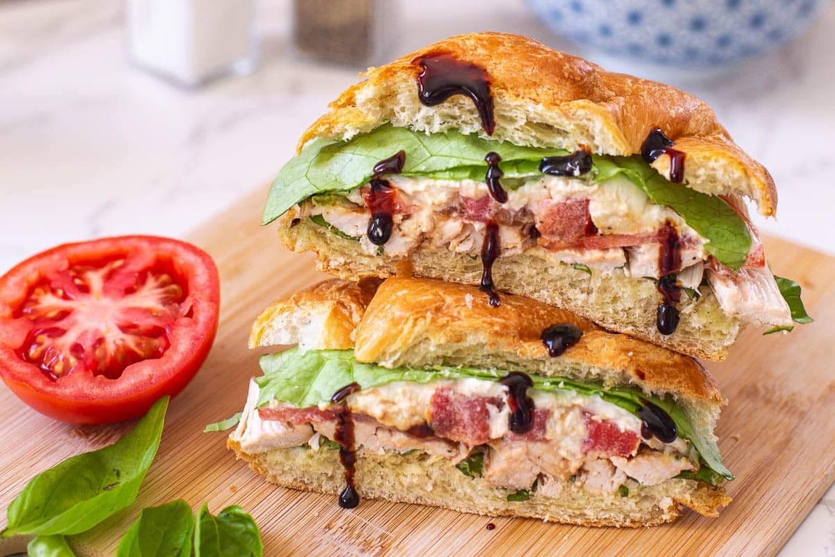 chicken croissant sandwich cut in half with balsamic glaze drizzled over it beside fresh tomato and basil