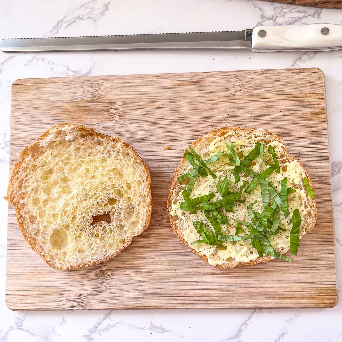 sliced croissant on cutting board with mayo and basil on one half beside bread knife