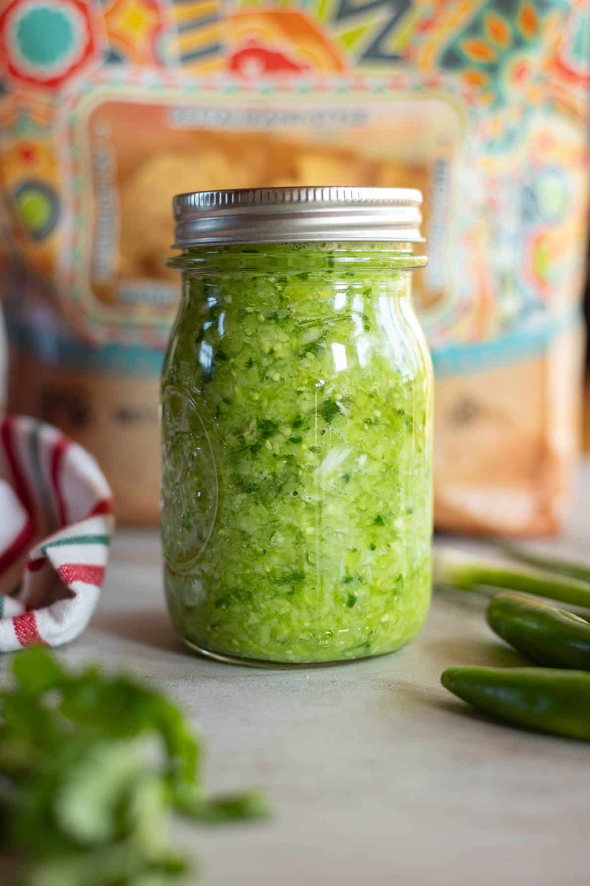 green tomato salsa in a jar with a bag of chips behind it.