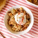 pear crumble in a small bowl with ice cream beside spoon, pears, and baking dish full of crumble