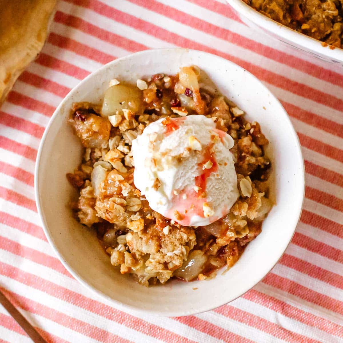 pear crumble in a small bowl with ice cream beside spoon, pears, and baking dish full of crumble