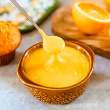 orange curd being drizzled from spoon into bowl beside muffin and sliced orange