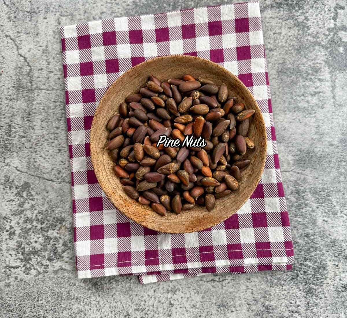 pine nuts in a wooden bowl on a checkered kitchen towel.