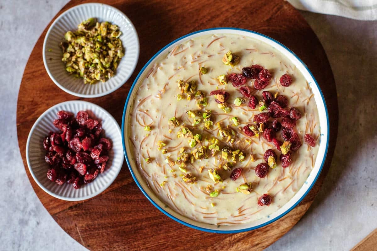 seviyan (sweet vermicelli dessert) in bowl with chopped pistachios and craisins on top and in small dishes off to the side.