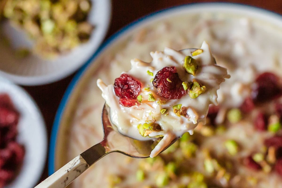 shemai in spoon above bowl with chopped pistachios and craisins on top and in small dishes off to the side.