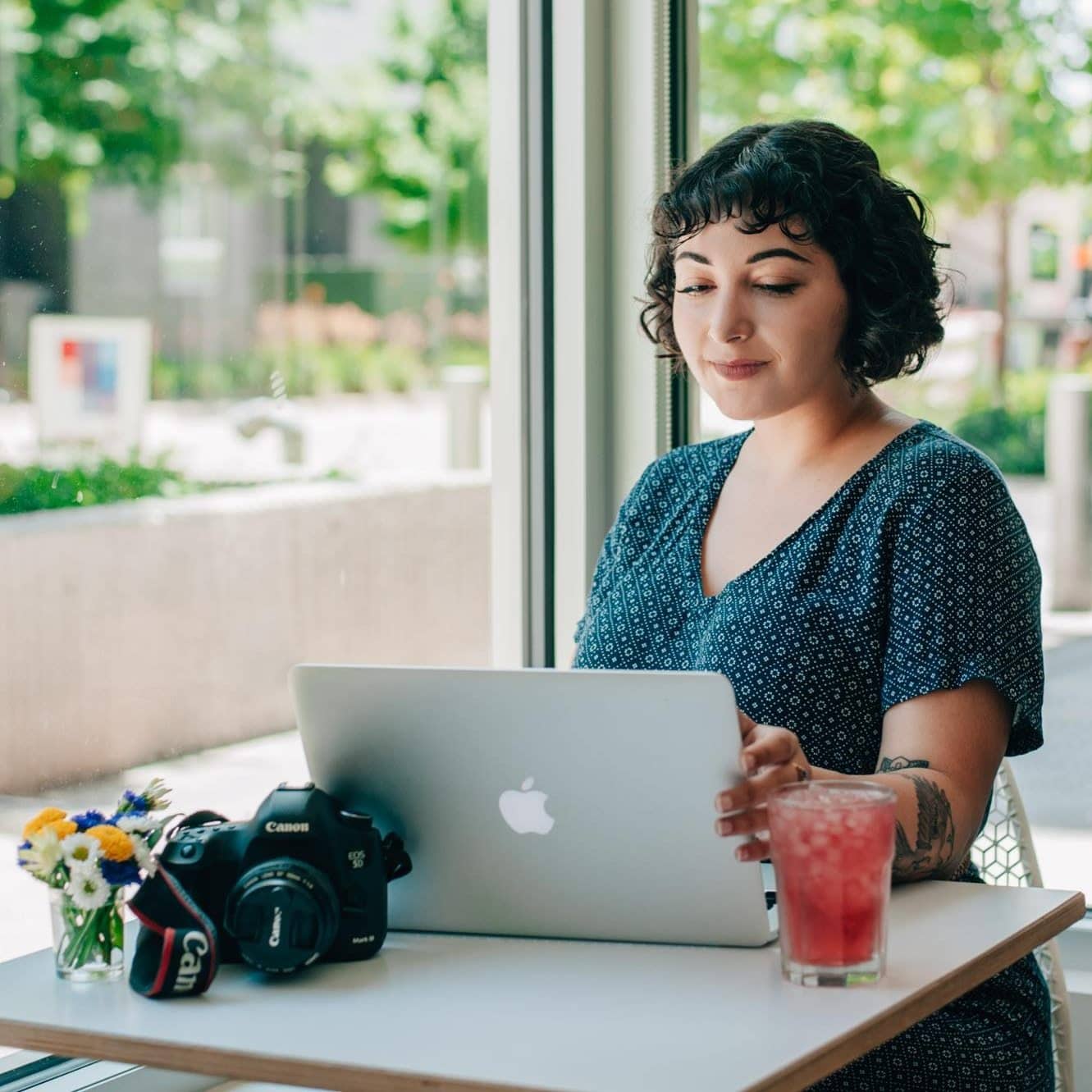 a lady sitting in a coffee shot wiht a pin drink, looking at her laptop with a camera on the table.