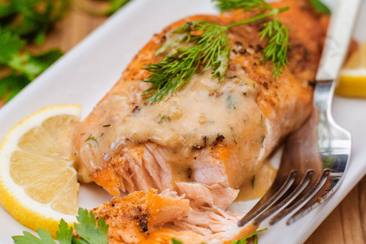 lemon and dill sauce drizzled over salmon beside herbs, lemon slices, fork.