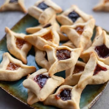 hamantaschen cookies on a blue plate with more cookies in the background.