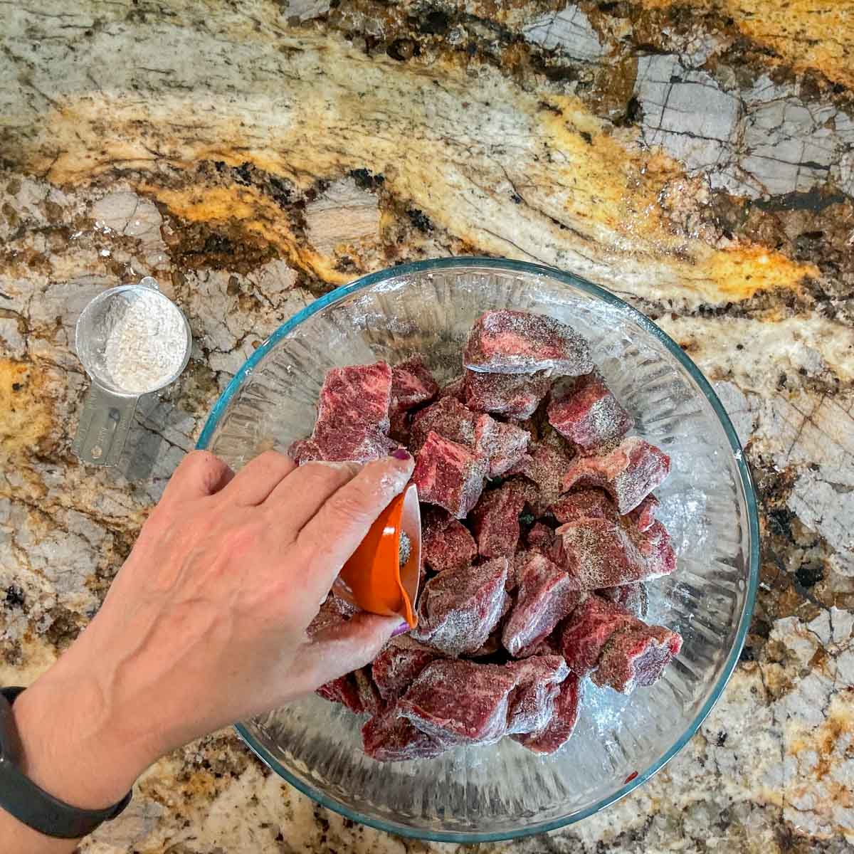 adding spices to a bowl of cubed raw meat. 