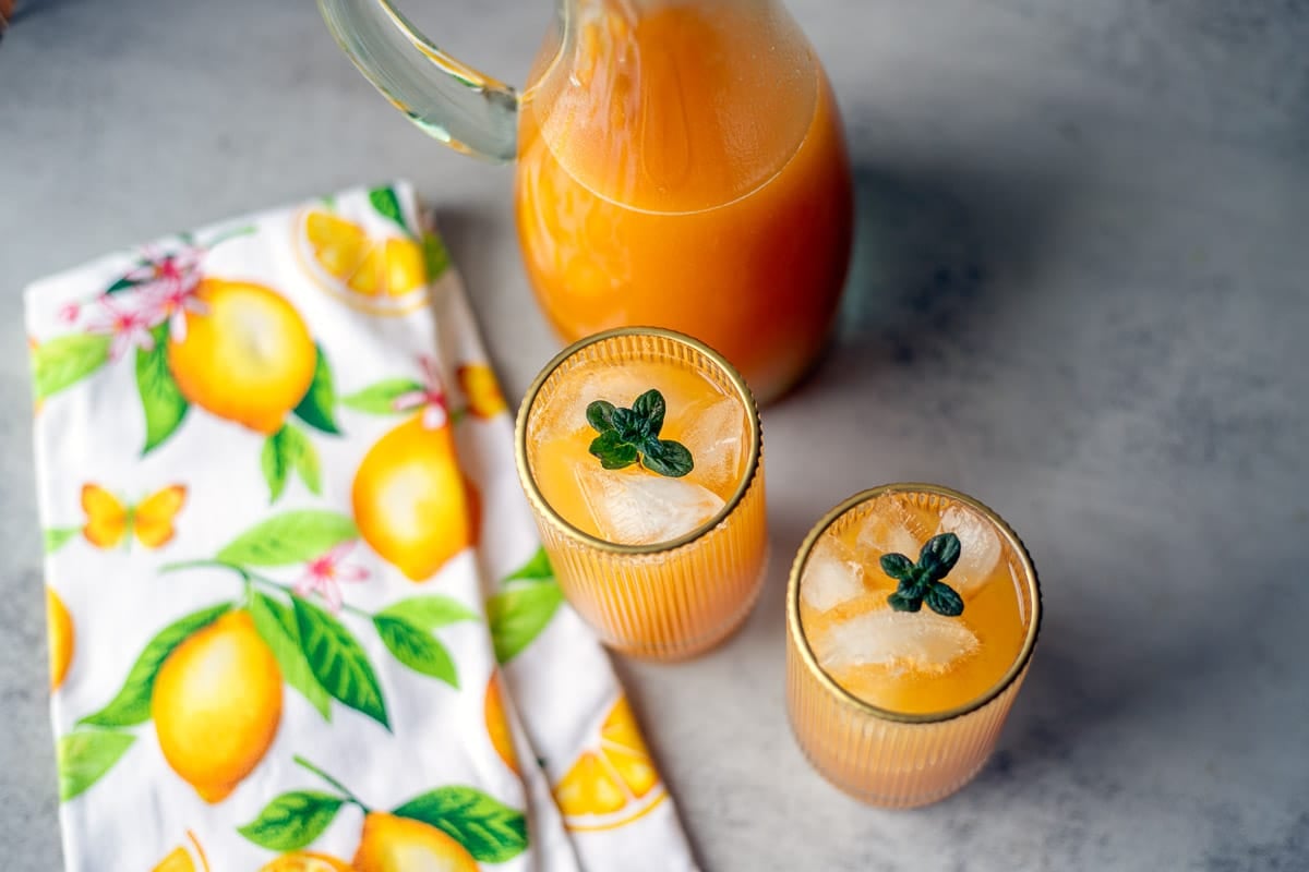 2 glasses with apricot juice, a lemon dish towel, and a pitcher. 