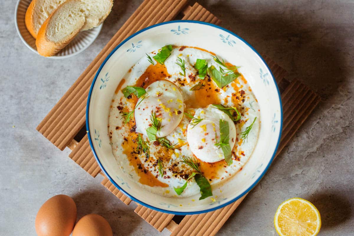 turkish eggs (cilbir) in a bowl with fresh herbs, sliced baguette, and lemon.