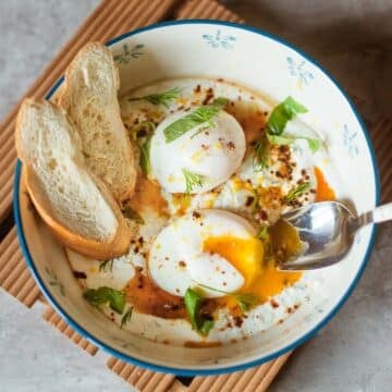 turkish eggs (cilbir) in a bowl with fresh herbs, sliced baguette, and spoon.