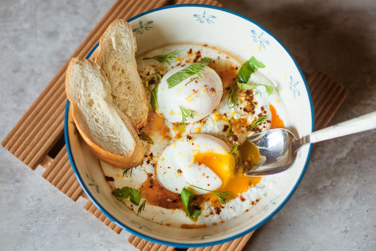 turkish eggs recipe (cilbir) in a bowl with fresh herbs, sliced baguette, and spoon.