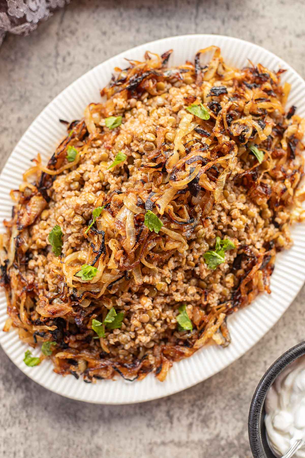 a white plate with Lebanese Mujadara  topped with caramelized onions and parsley, with a side of yogurt.