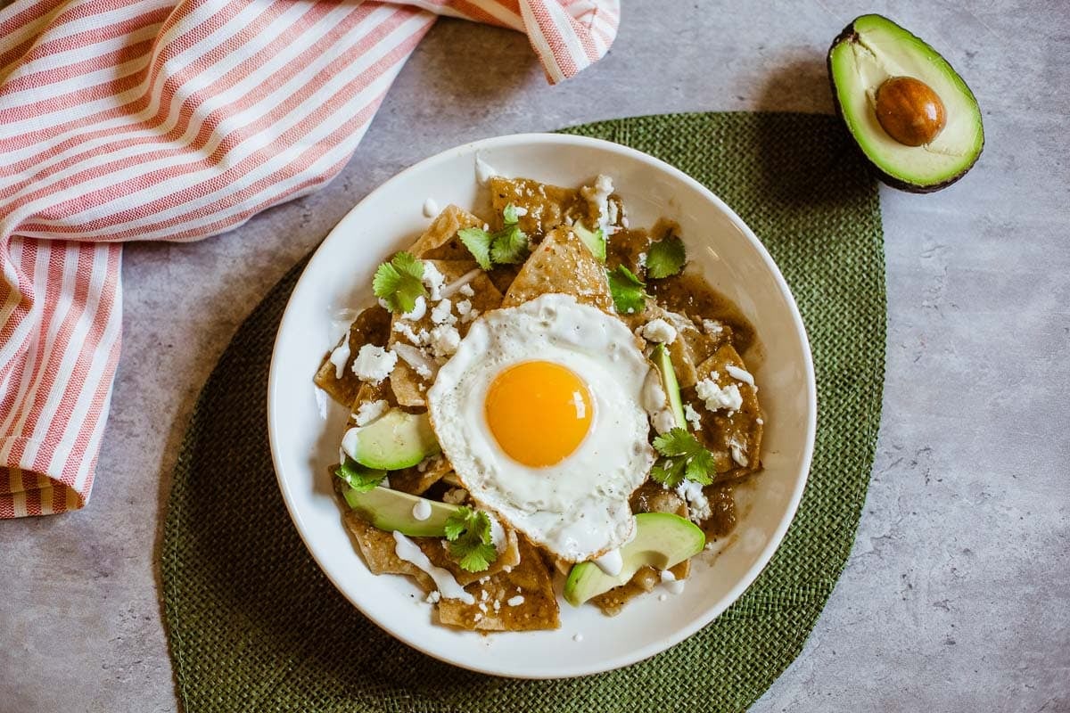 Chilaquiles verde with an egg on top on a green mat and ½ of an avocado next to it.