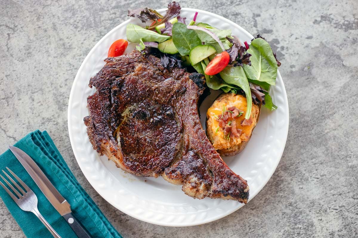 smoked ribeye steak on a white plate with a baked potato and a salad.