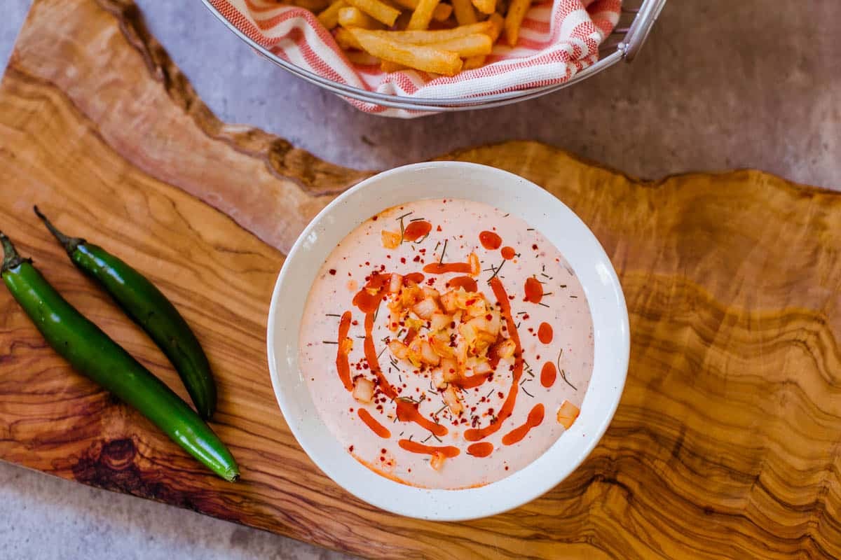 bowl of spicy ranch dip with a swirl of hot sauce and diced kimchi on top and fries and serranos on the side.