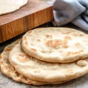 Greek pita bread stacked on one another next to a cutting board and a blue dish towel.