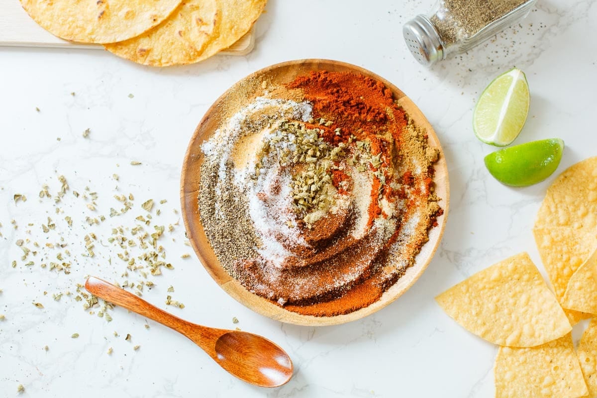 chicken taco seasoning on a plate after being partially mixed, surrounded by tortillas, spoon, lime, spices.