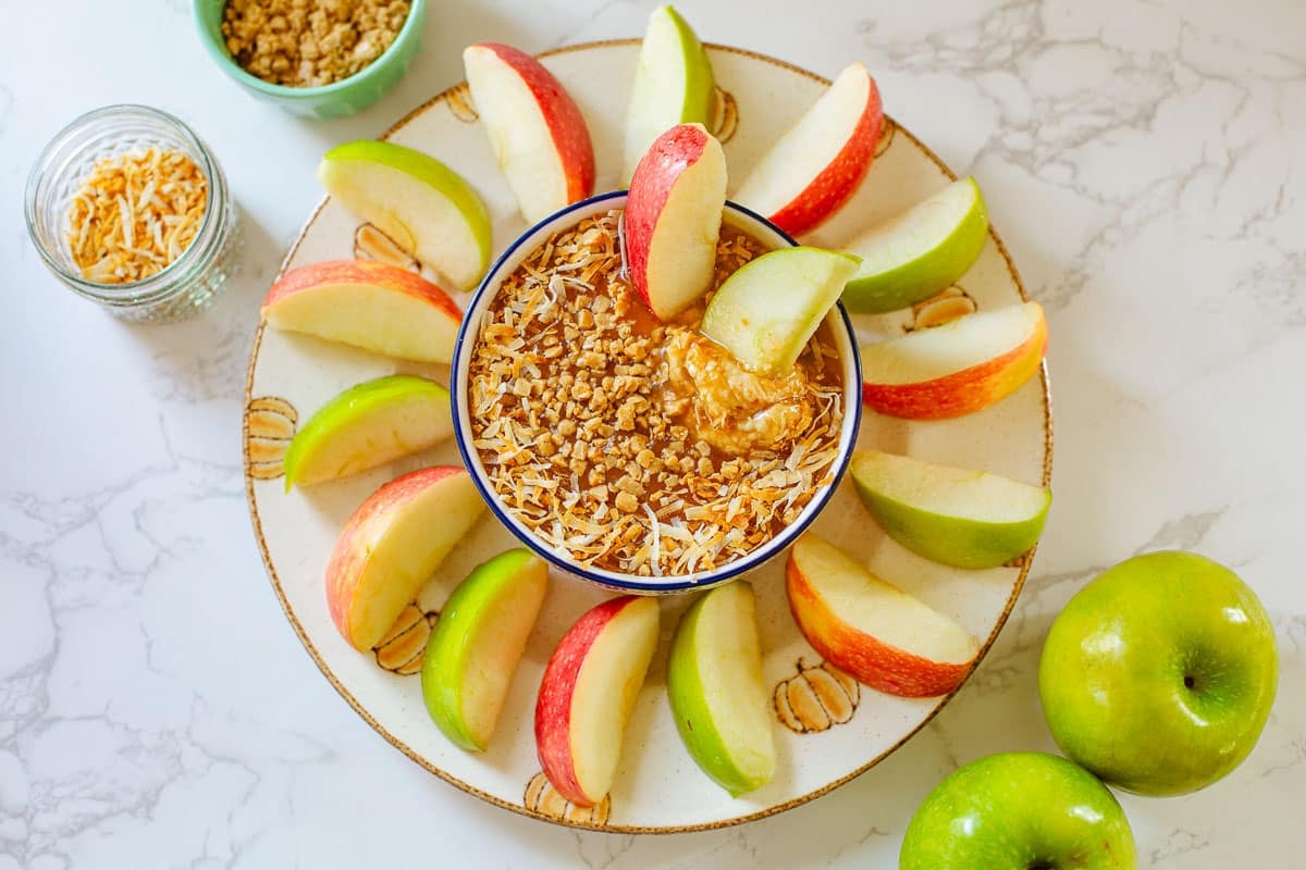 cream cheese caramel apple dip surrounded by apple slices, whole apples, toffee bits, and coconut flakes.